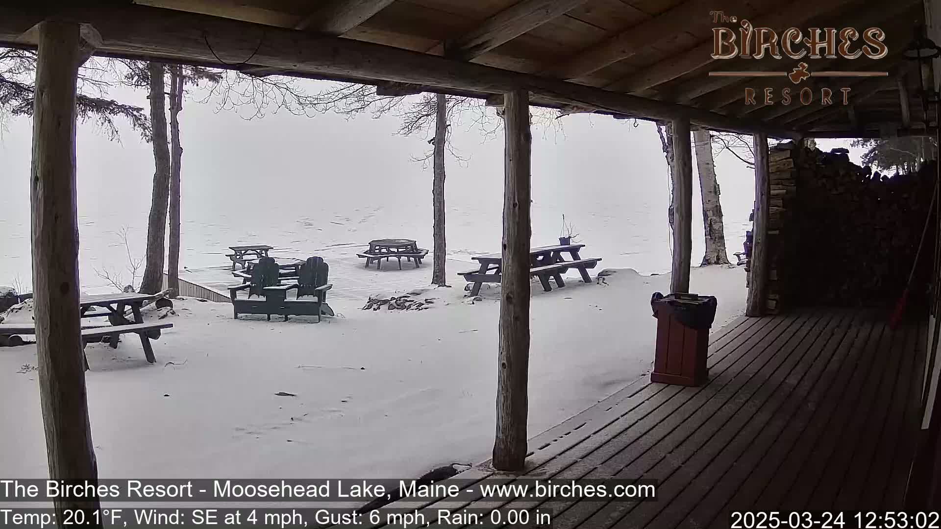 A snow-covered outdoor patio with picnic tables and Adirondack chairs overlooks a calm, snowy lake under an overcast sky.