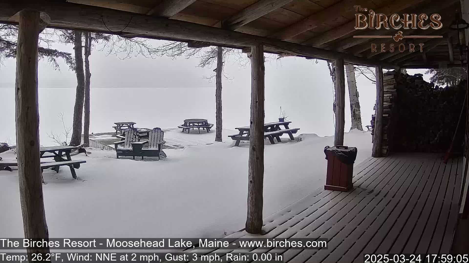 A snow-covered porch overlooks a lake, with several picnic tables and chairs scattered in the snow.