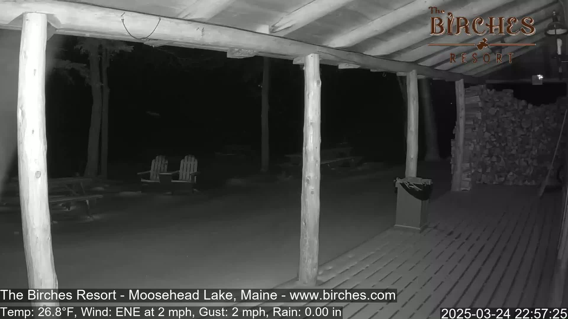 A nighttime view from a porch shows a snow-covered deck, two Adirondack chairs, a woodpile, and a trash can under a covered porch with wooden posts, in rainy conditions with a light east-northeast wind.