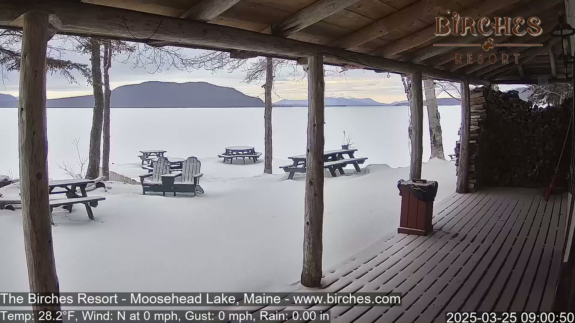 A snow-covered lakeside patio with picnic tables and Adirondack chairs is viewed from a wooden porch, mountains and a frozen lake visible in the background under a cloudy sky.