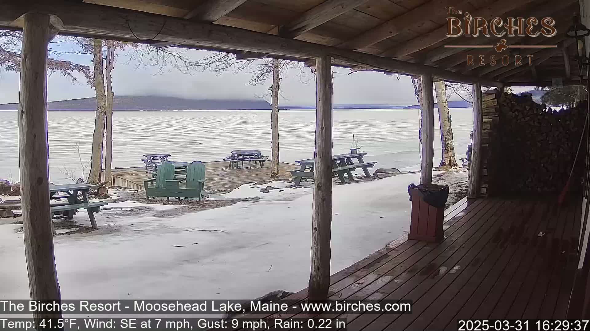 A partially frozen lake is viewed from a wooden porch, with picnic tables and Adirondack chairs on a snow-covered dock under a cloudy, rainy sky.