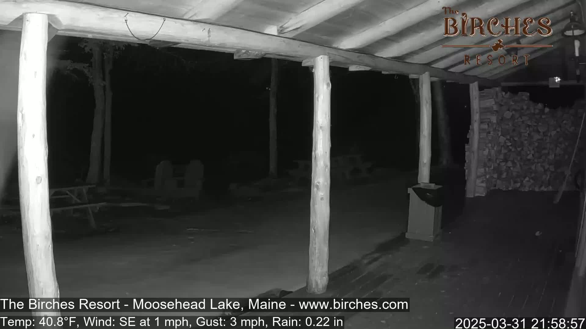 A nighttime, rainy view from a covered porch shows a dark outdoor area with wooden posts, a pile of firewood, and some indistinct furniture.