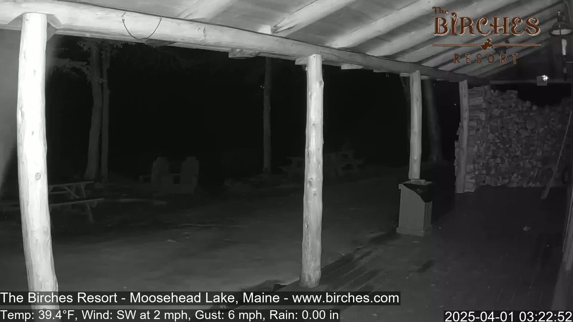 A nighttime view from under a covered porch shows a dark outdoor area with picnic tables and a woodpile, under a southwest wind with light rain.