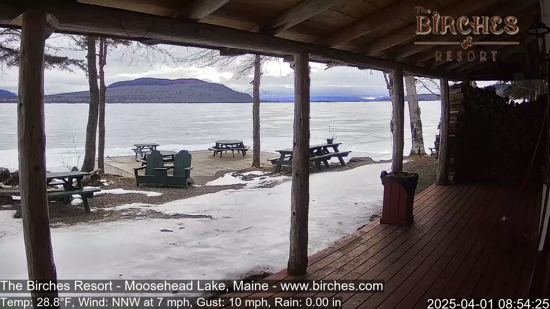 A partially frozen lake is viewed from a wooden porch with picnic tables and Adirondack chairs scattered across a snow-covered patio, under a cloudy sky.