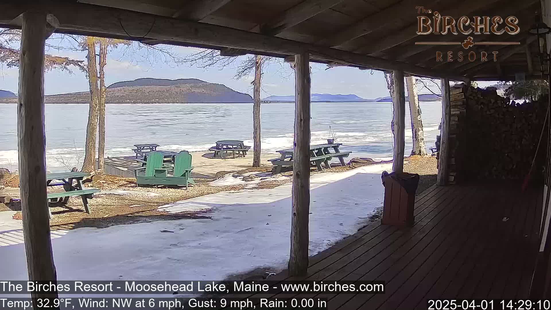 A partially frozen lake is viewed from a wooden porch on a sunny day with snow on the ground and picnic tables outside.