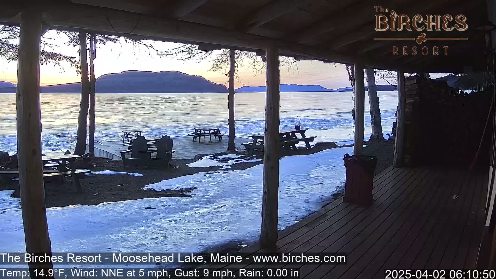 A view from a lakeside porch shows a partially frozen lake at sunset with picnic tables and a dock, surrounded by snow and bare trees under a calm sky.
