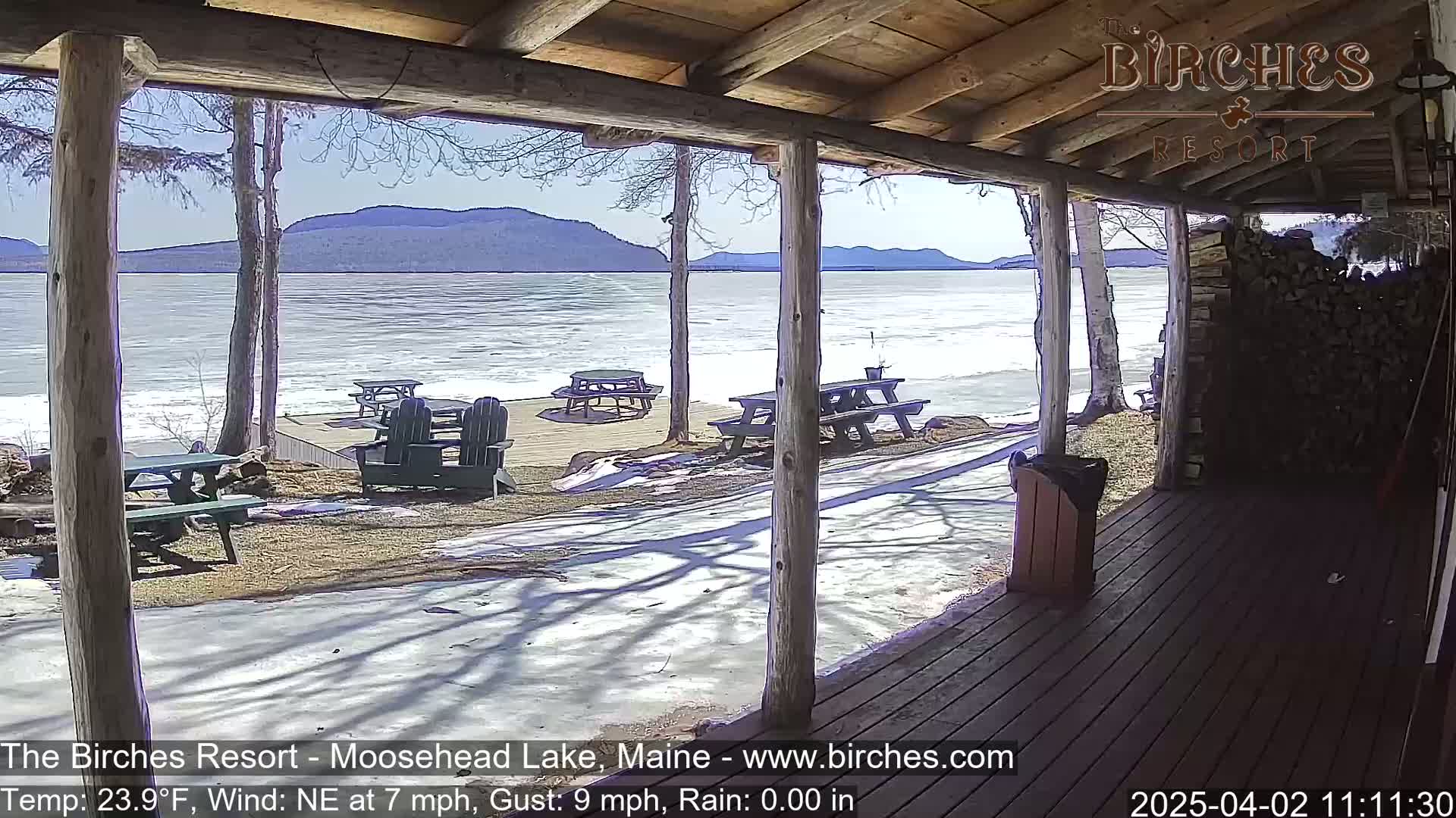 A partially covered porch overlooks a partially frozen lake with mountains in the background on a sunny, windy day.