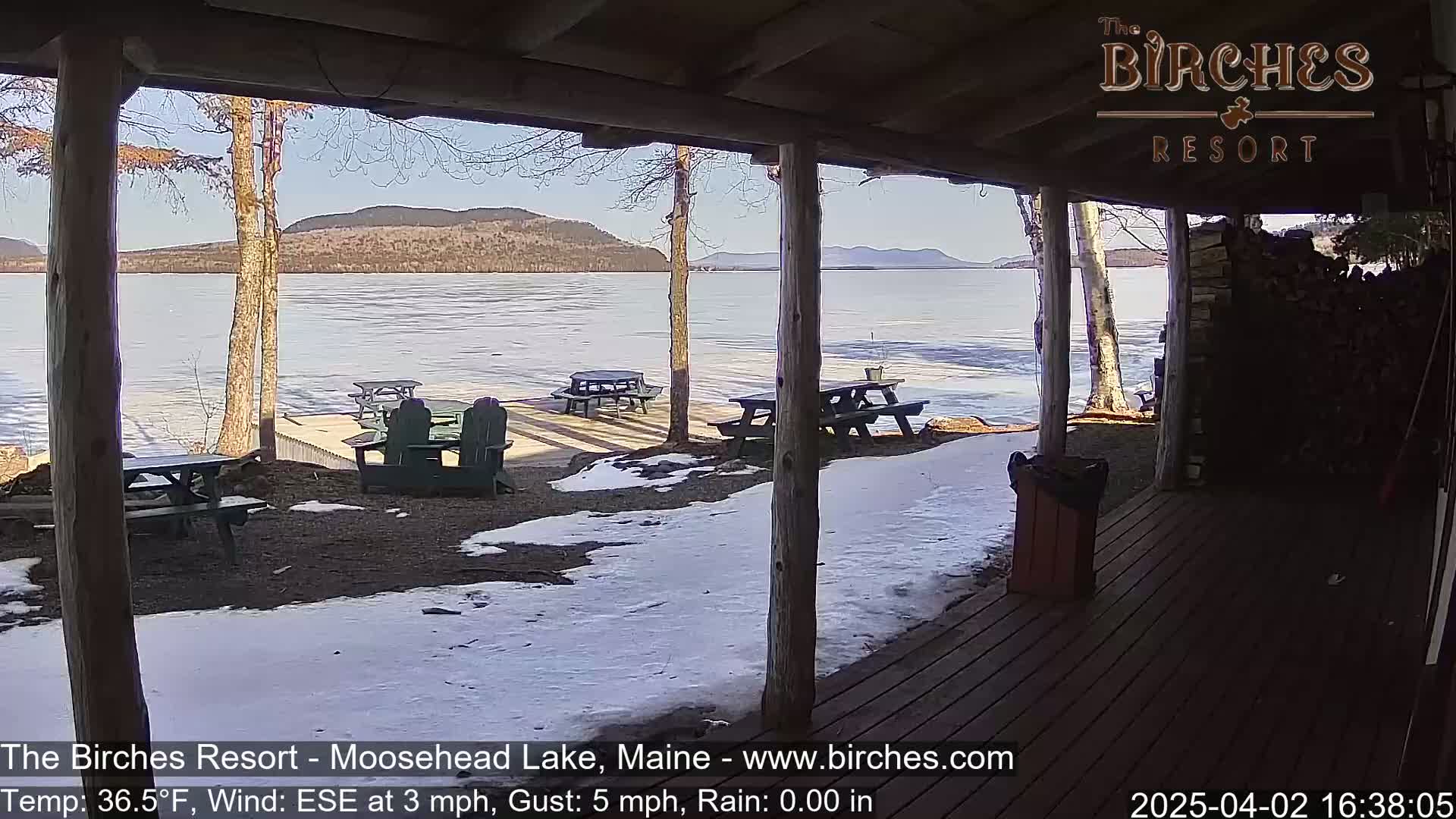 A partially frozen lake is visible from a wooden porch on a sunny day with some snow on the ground.