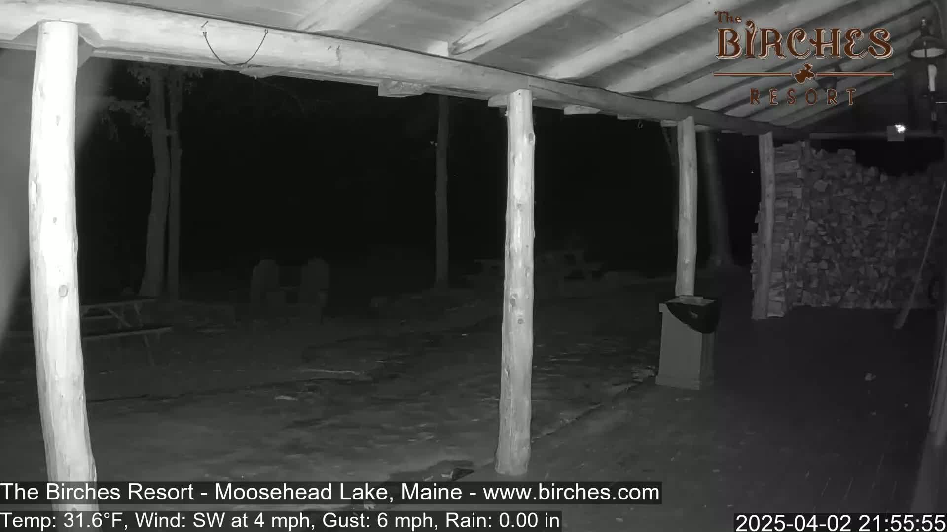 A nighttime, black and white view of a covered patio area with wooden posts and a pile of firewood, under a southwest wind of 4 mph with gusts up to 6 mph, and no rain.