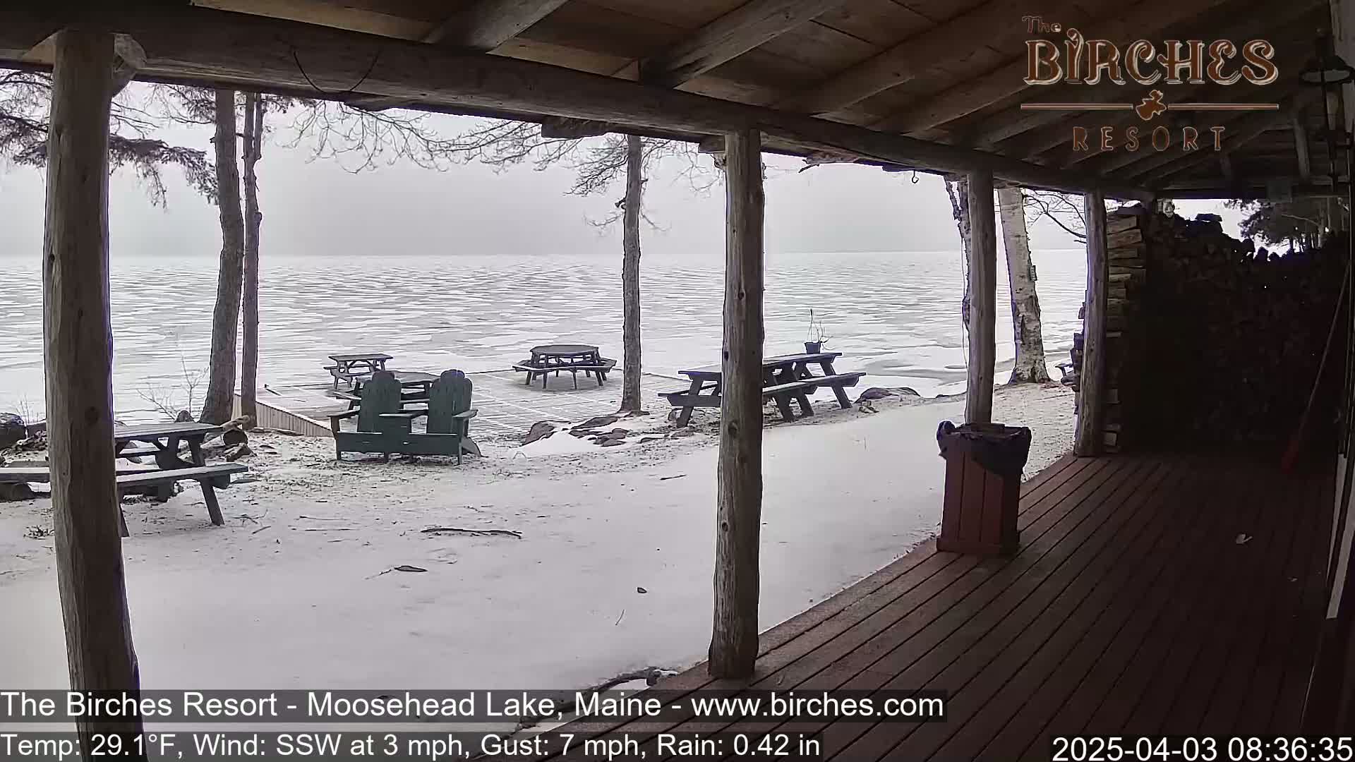 A snow-covered lakeside patio with picnic tables is viewed from a porch during rainy, windy weather.