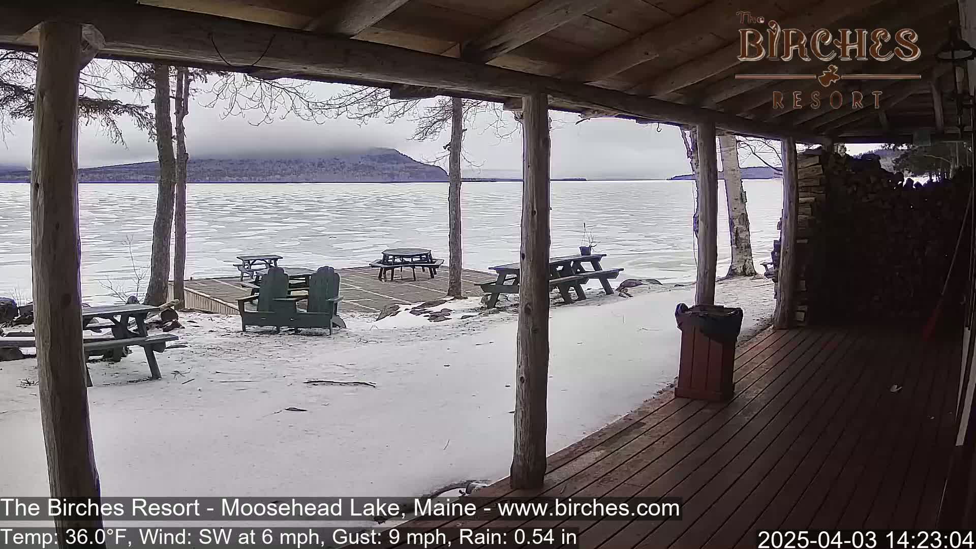 A snow-covered lakeside patio with picnic tables and chairs is viewed from a wooden porch under an overcast sky.