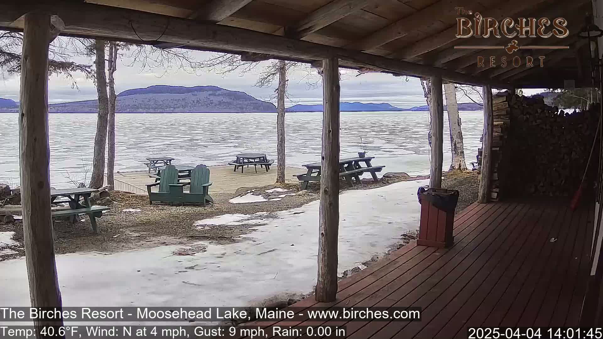A partially frozen lake is viewed from a wooden porch with picnic tables and Adirondack chairs, under a cloudy sky.