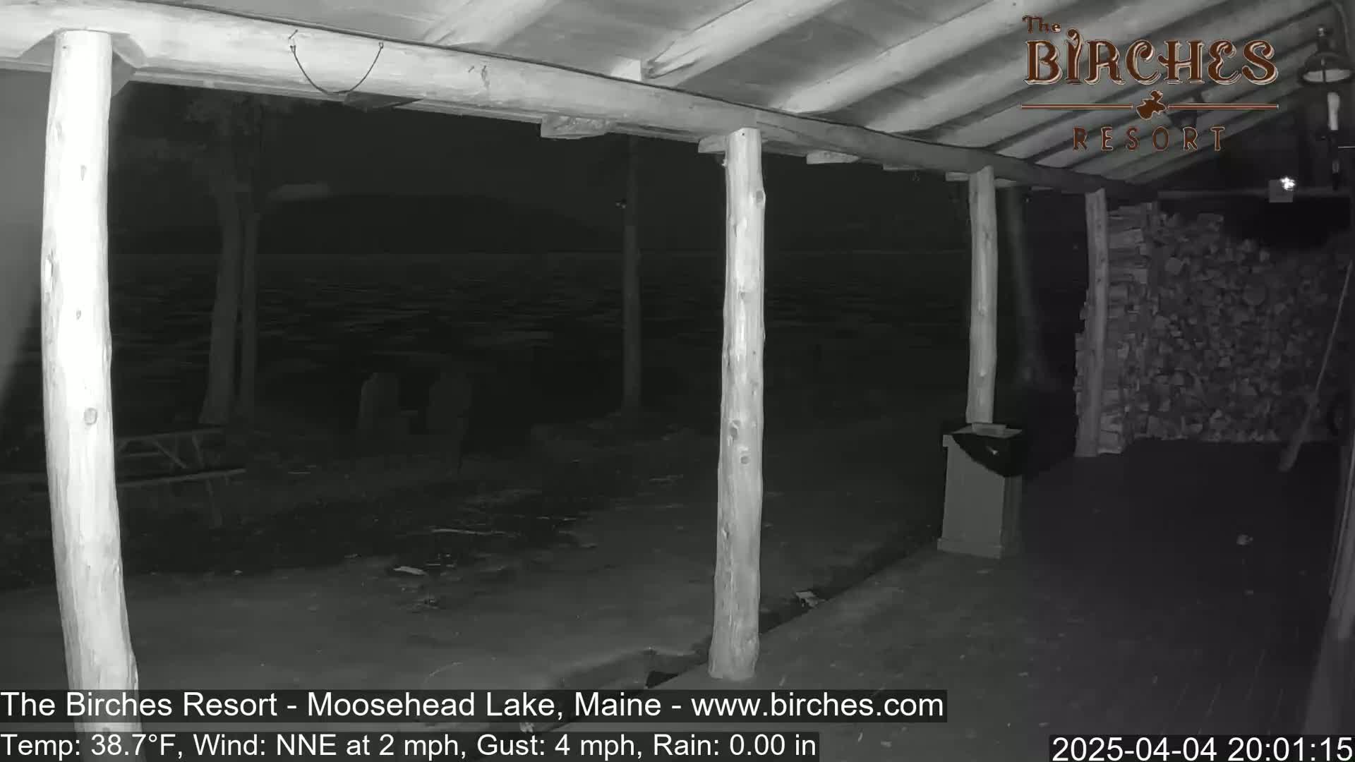 A nighttime, low-light view from under a covered porch shows a dark lake, trees, and a picnic table in the distance, with a woodpile visible to the right, under conditions of a 38.7°F temperature, a NNE wind at 2 mph with gusts up to 4 mph, and no rain.