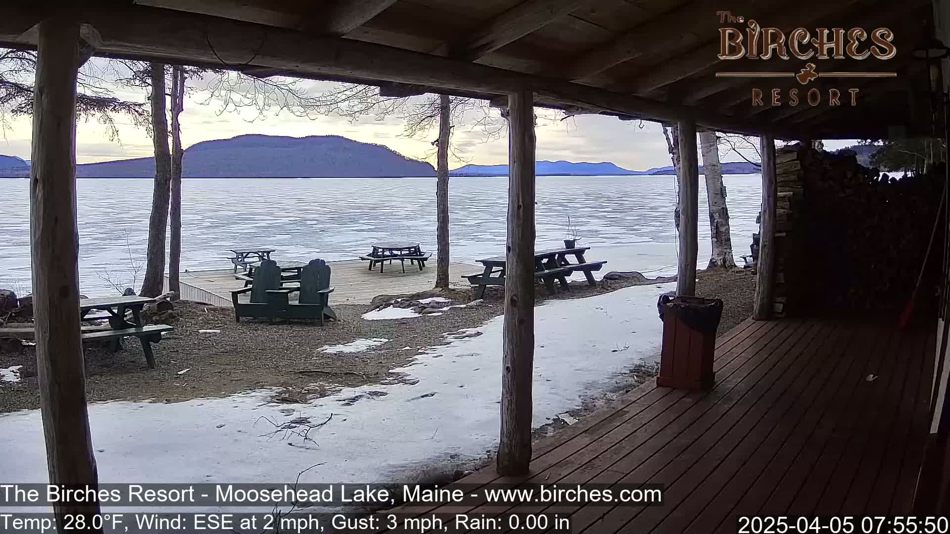 From a wooden porch overlooking a partially frozen lake, several picnic tables and Adirondack chairs sit on a snow-dusted deck under a cloudy sky, with distant mountains visible.