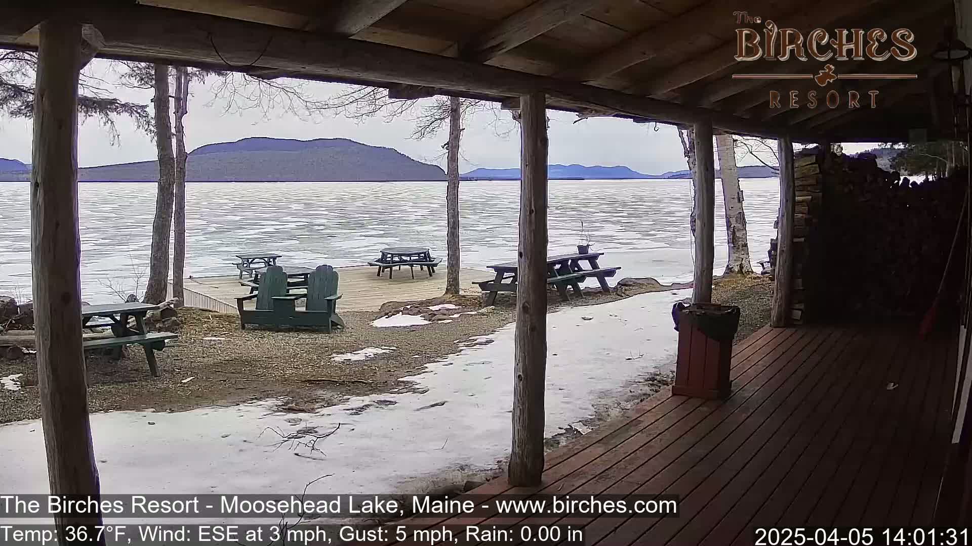 A partially frozen lake is viewed from a wooden porch with picnic tables and Adirondack chairs, under a cloudy sky with light snow on the ground.