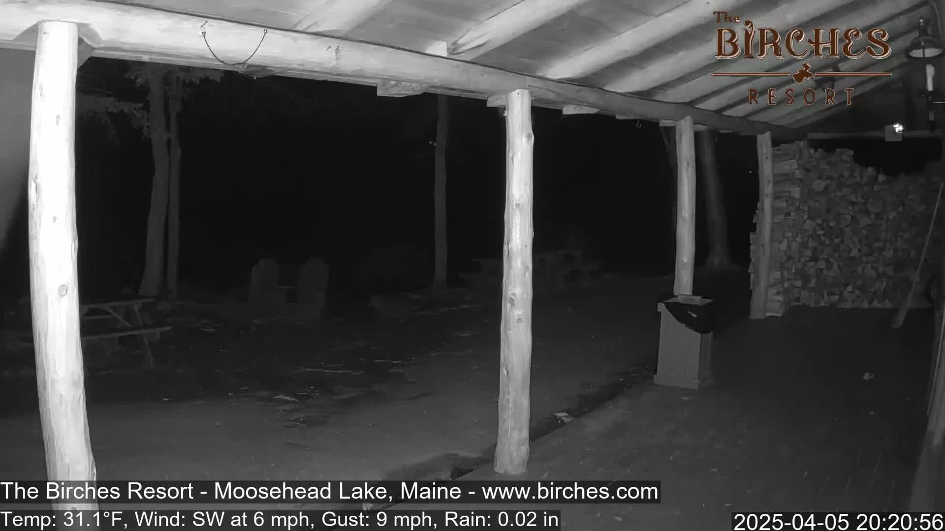 A nighttime, rainy view of a covered porch area with wooden posts and a woodpile, under a dark sky.