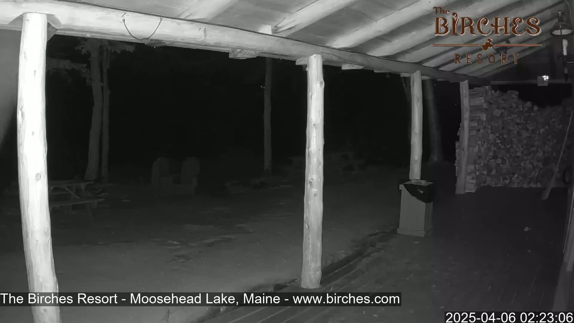 A nighttime view of a covered porch with wooden posts, a picnic table in the background, and a stack of firewood to the right.