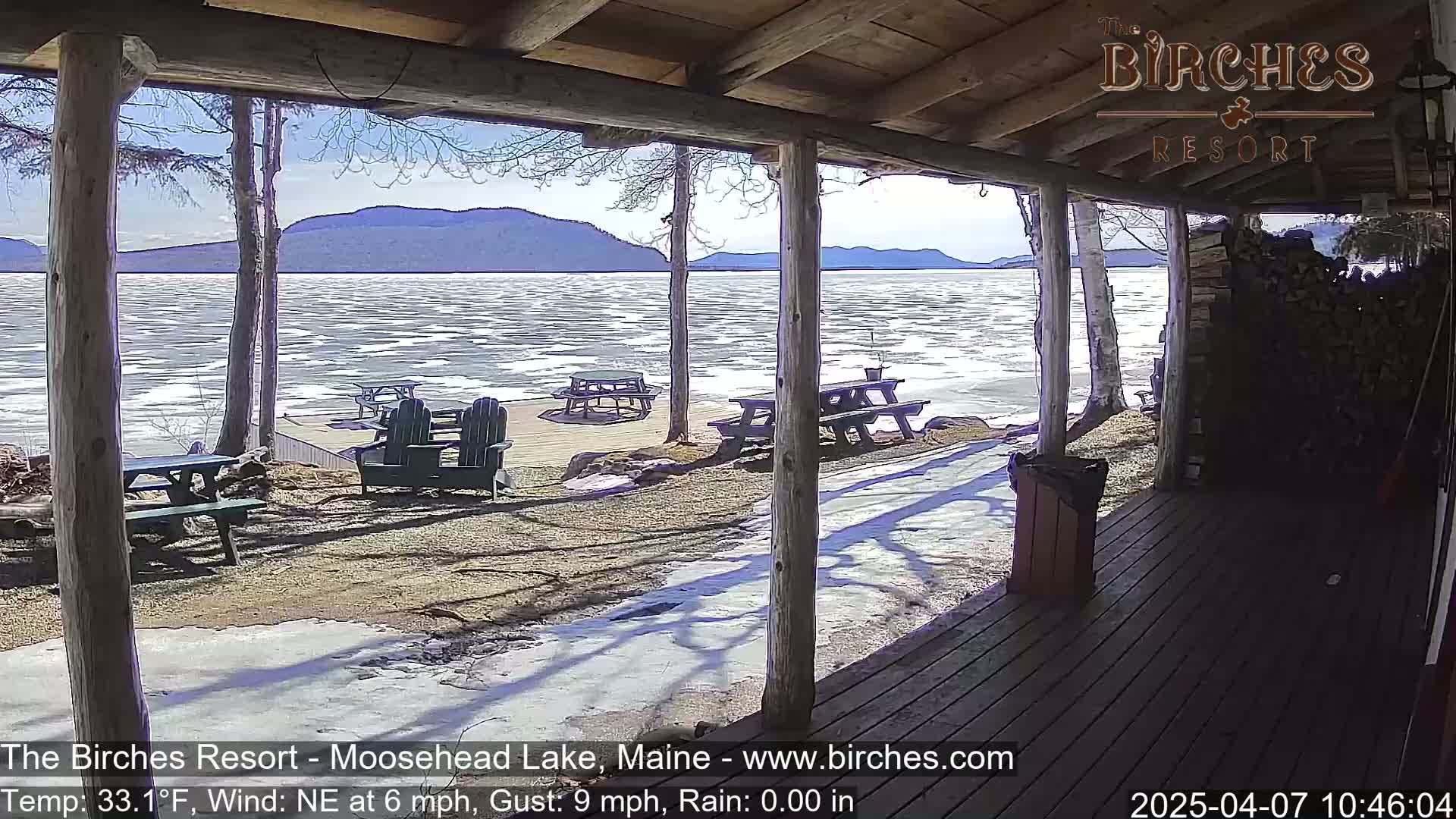 From a porch overlooking a partly frozen lake and distant mountains, several picnic tables and Adirondack chairs sit on a partially snow-covered ground under a sunny sky.
