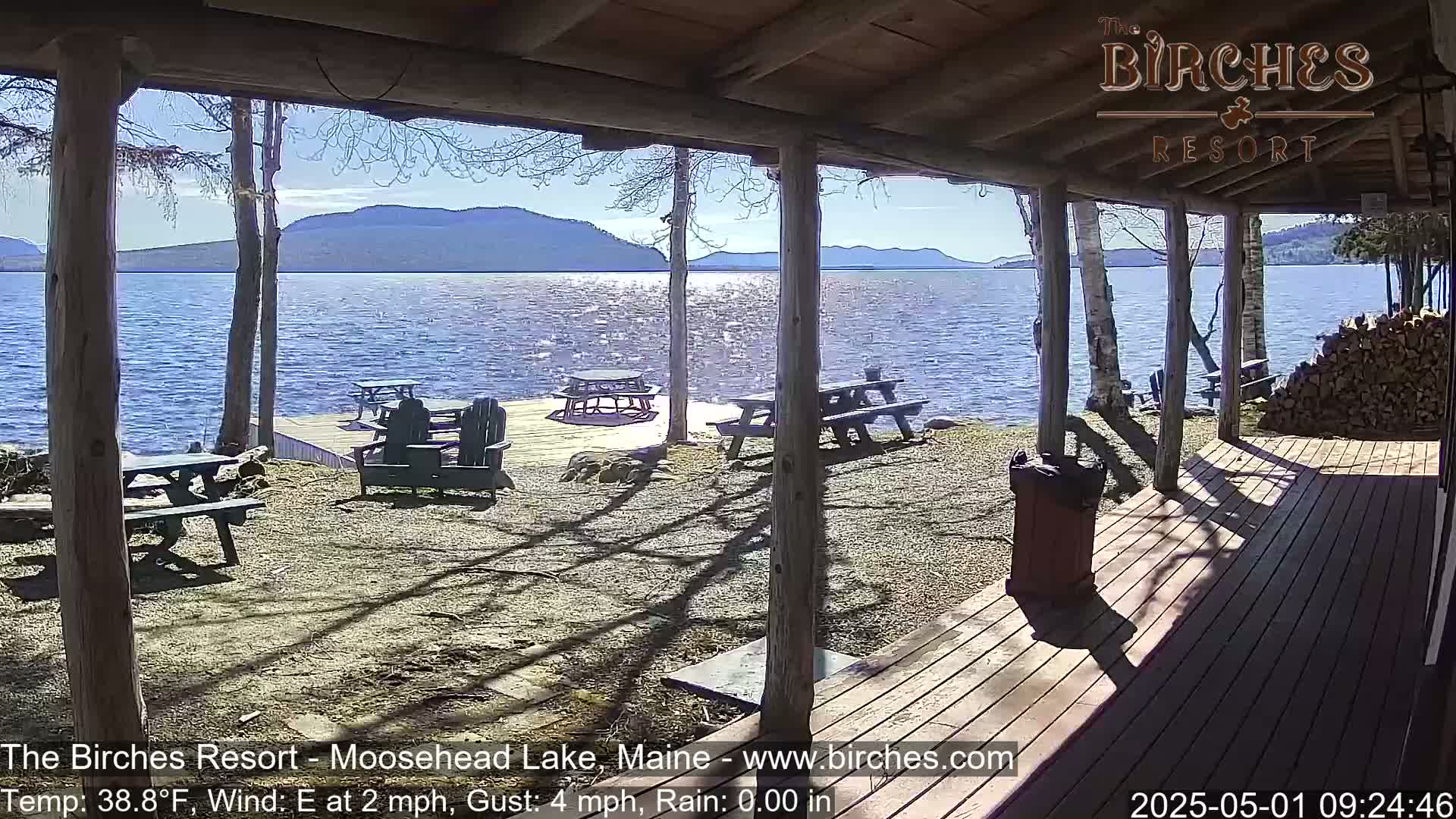 A sunny view of a lake and mountains from a lakeside porch with picnic tables and chairs is shown.