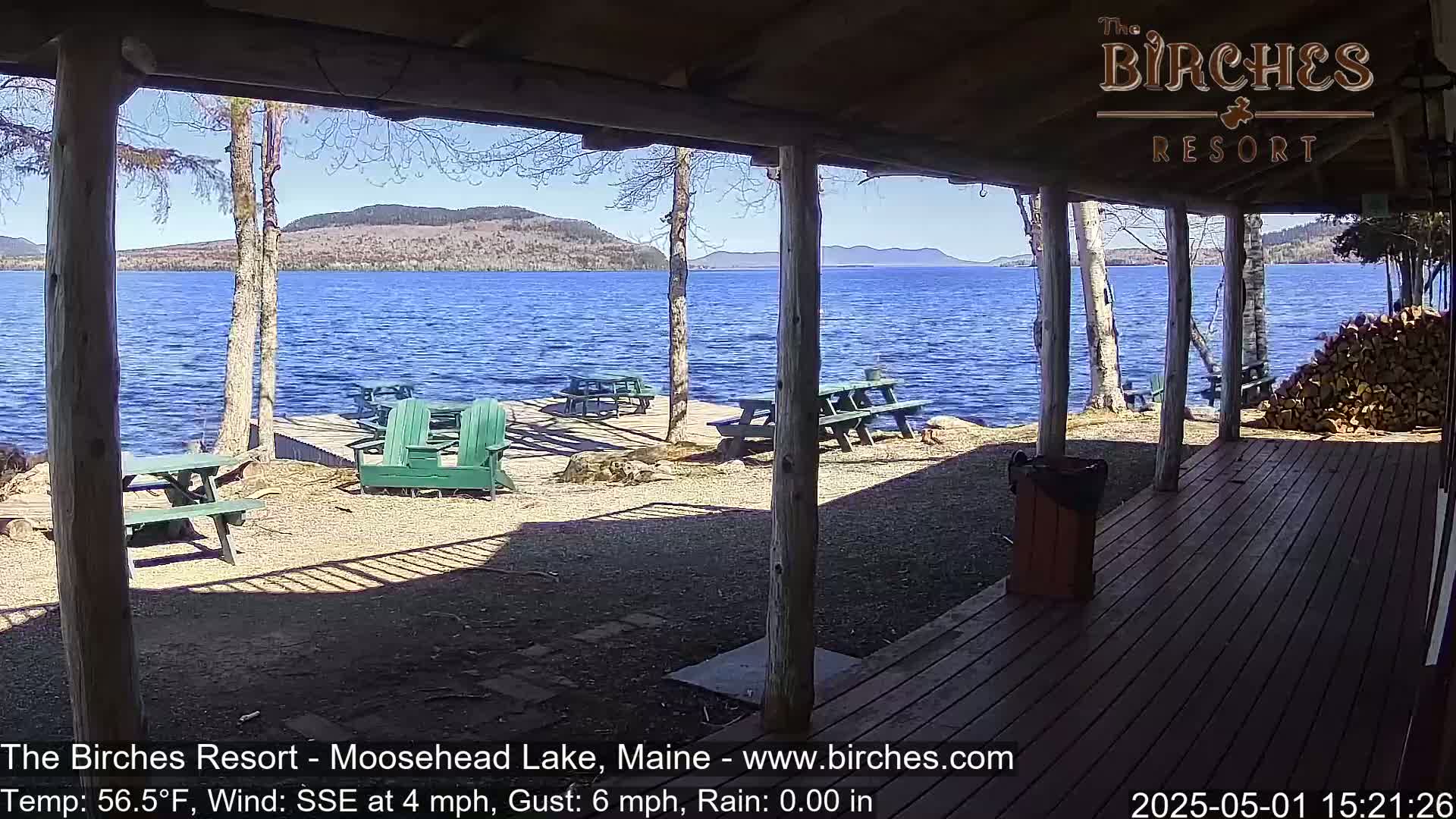 A sunny view from a porch overlooking a calm lake with picnic tables and Adirondack chairs on a gravel area, backed by trees and distant mountains.