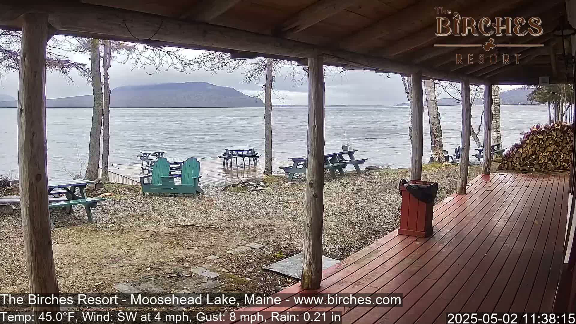 A rain-swept lakeside view from a wooden porch shows picnic tables and Adirondack chairs scattered on a gravel area, with a calm lake and distant mountains under an overcast sky.