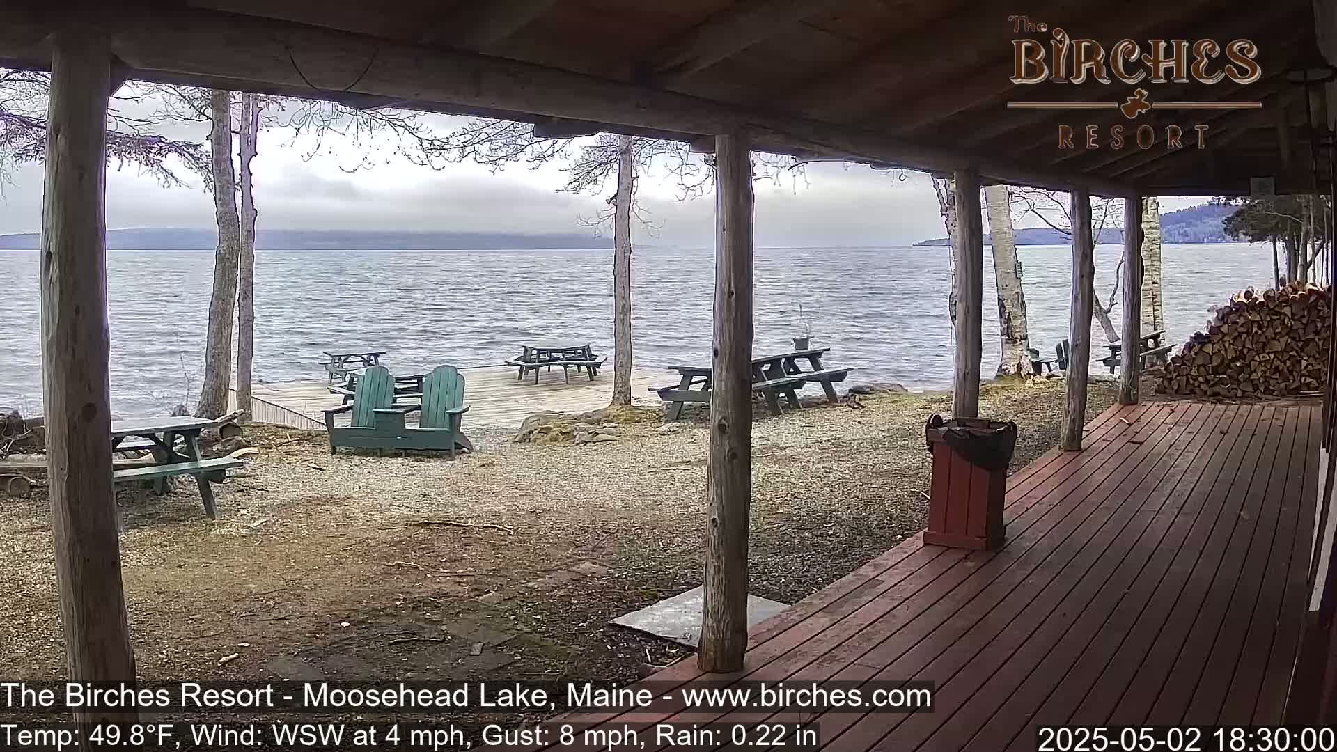 A lakeside view from a covered porch shows several picnic tables and Adirondack chairs on a gravel area leading to a wooden dock, with a calm lake under an overcast sky.