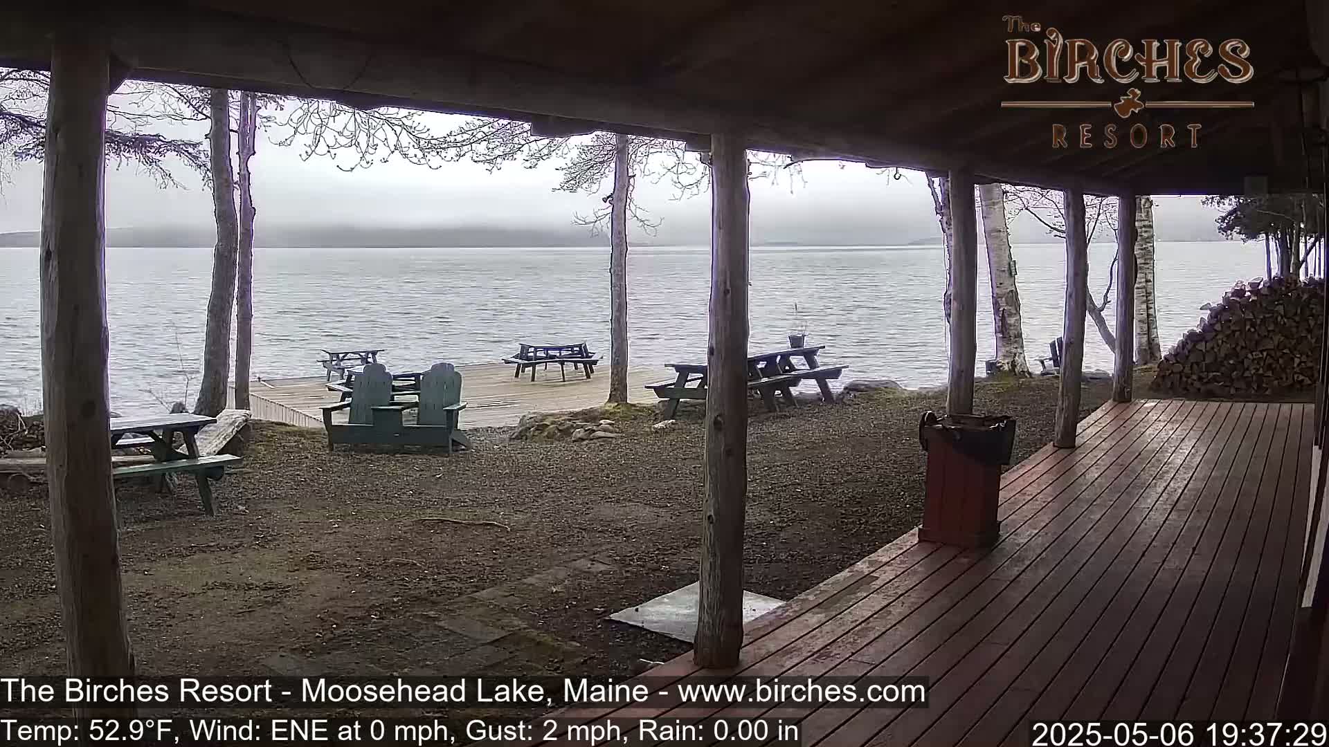 A view from a covered porch shows a calm lake with several picnic tables and chairs on a dock in rainy conditions.