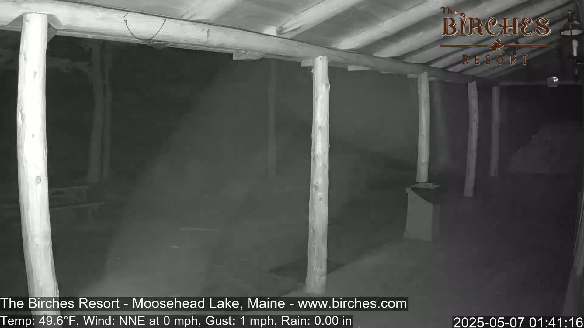 A nighttime, rainy scene shows a covered outdoor area with wooden posts and a hazy atmosphere.