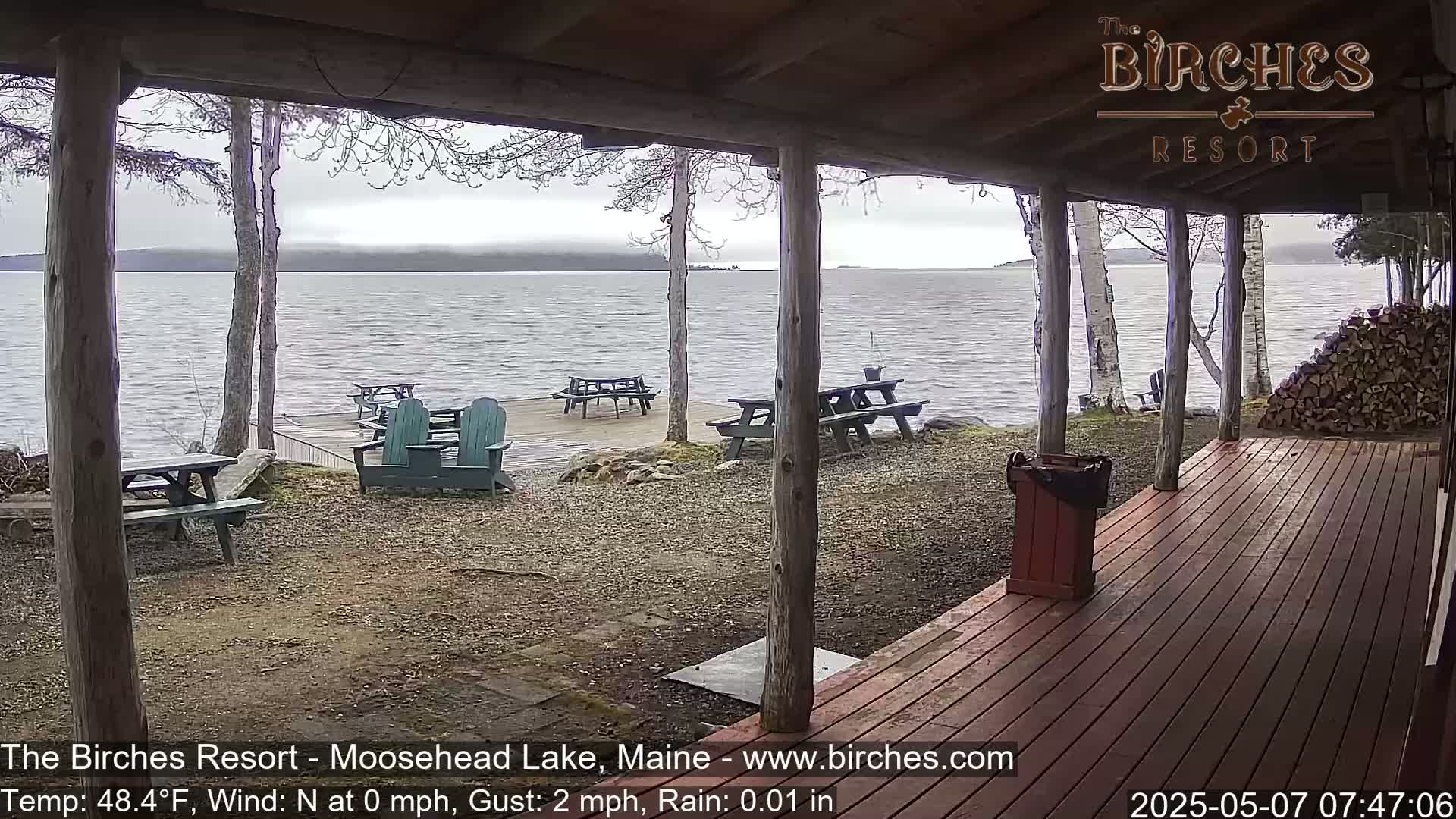 A covered porch overlooks a calm lake with picnic tables and Adirondack chairs on a gravelly shore under an overcast sky with light rain.