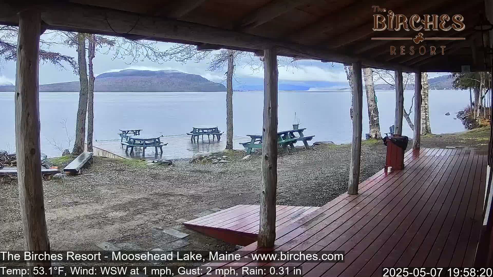 A view from a covered porch shows a calm lake with several picnic tables on a wet dock, under an overcast sky with light rain.