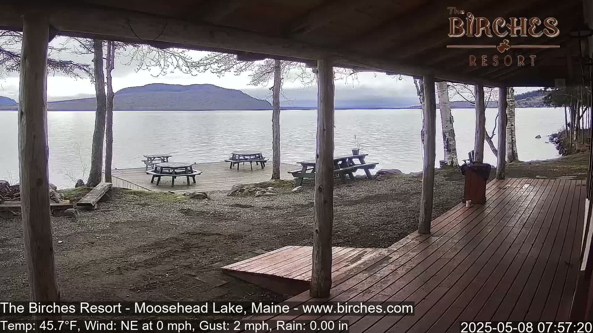 From a covered porch, several picnic tables are visible on a dock overlooking a calm lake with mountains in the background, under an overcast sky.
