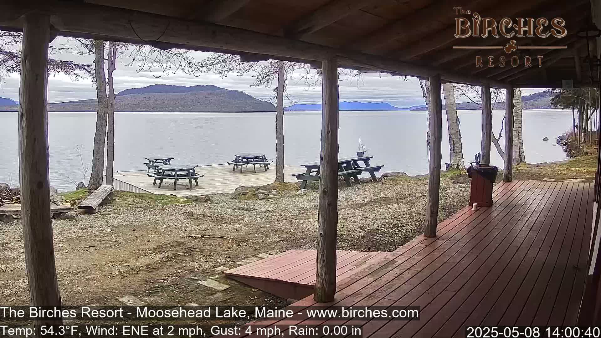 A covered porch overlooks a calm lake with several picnic tables on a dock in the background under a cloudy sky.