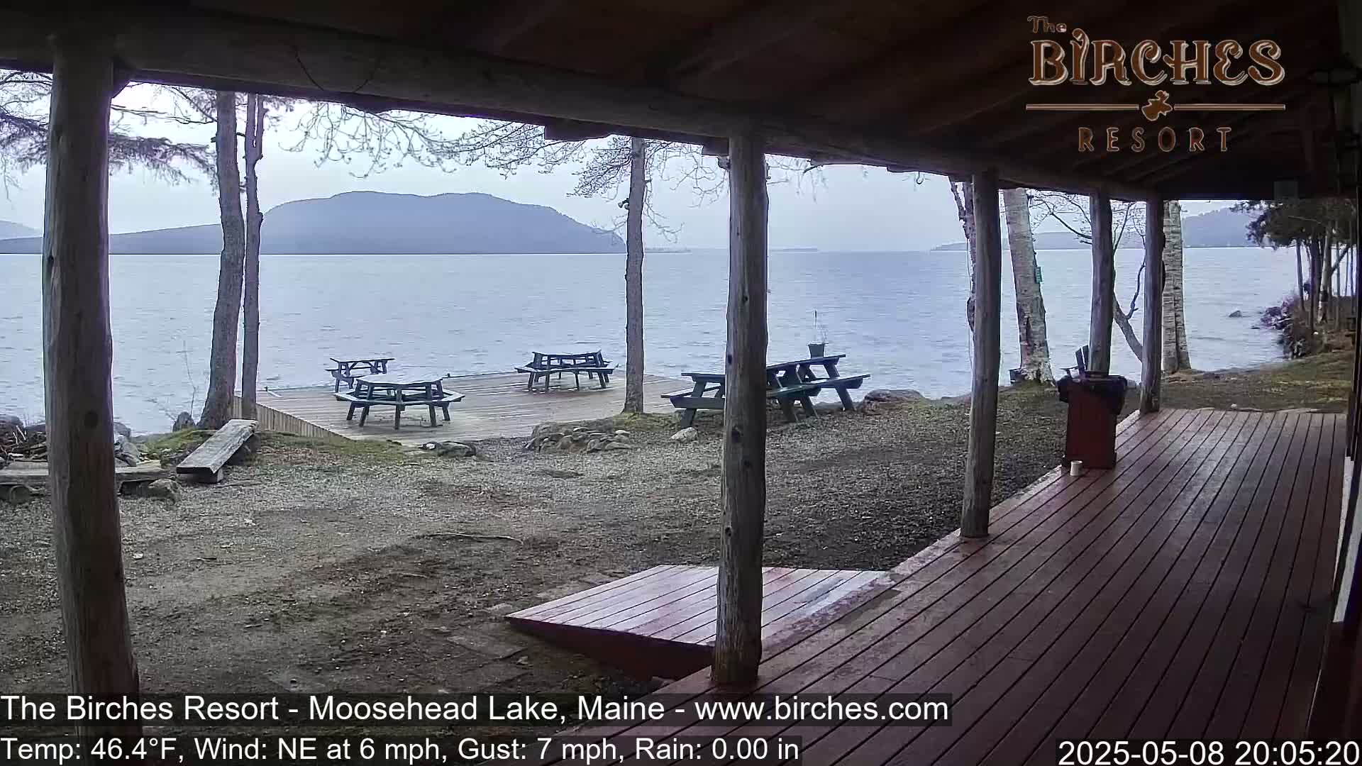 A covered porch overlooks a calm lake with several picnic tables on a dock in the rain.