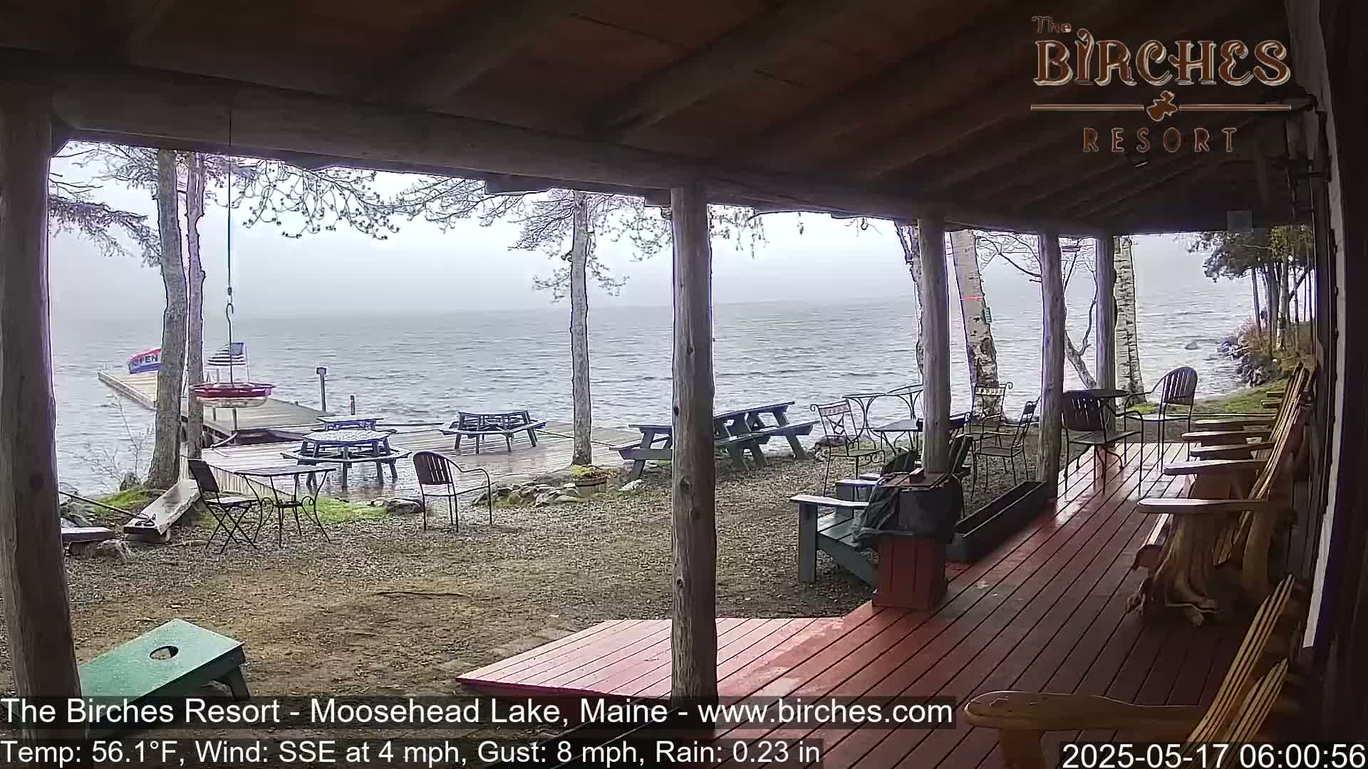 A rain-misted lake is viewed from a covered porch with wooden furniture and a small dock in the distance.