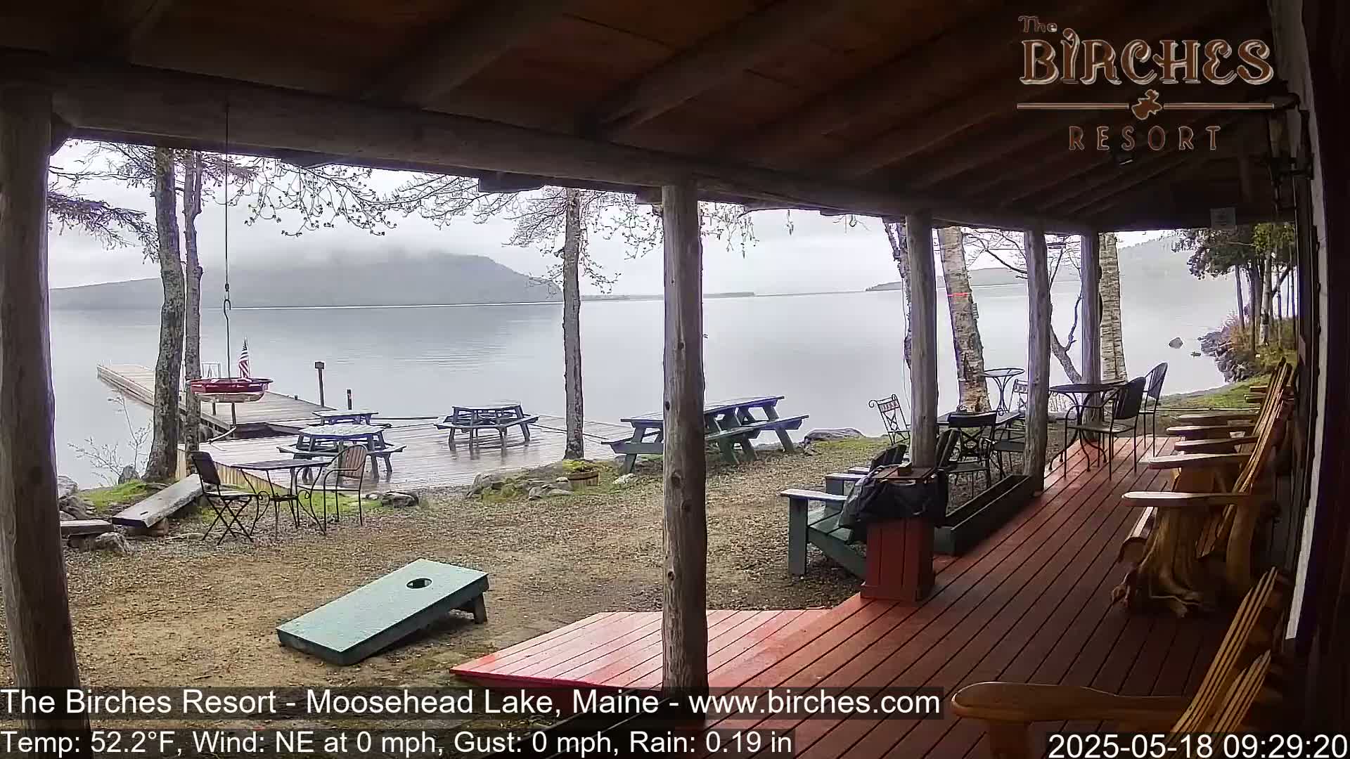 A rainy lakeside porch with picnic tables, Adirondack chairs, and a cornhole game, overlooking calm water and a distant mountain.