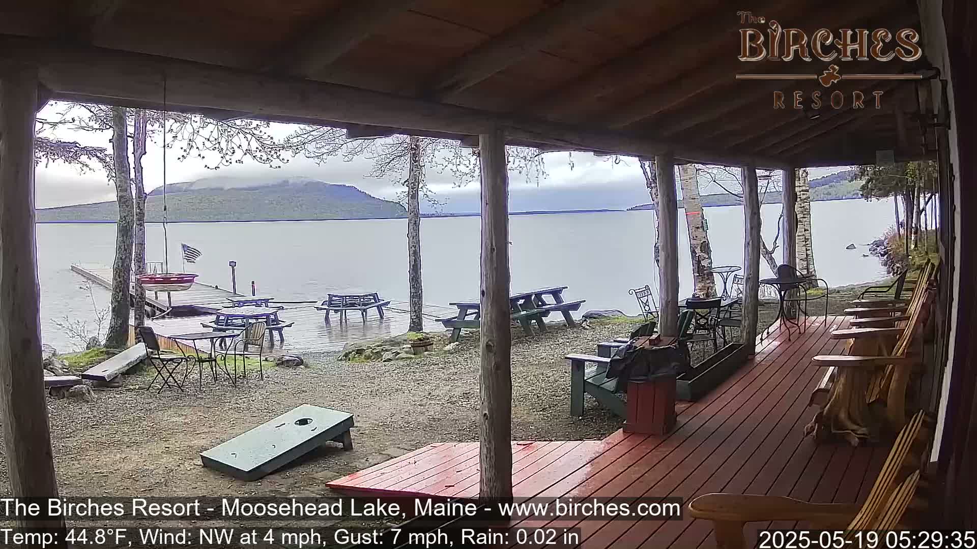 A lakeside porch with several picnic tables and Adirondack chairs overlooks a calm lake under a cloudy, rainy sky.