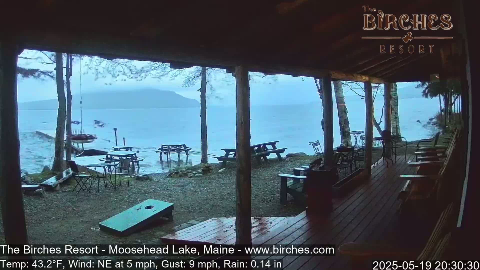 A rainy view from a covered porch shows a lake with a distant mountain under a gray sky, several picnic tables and chairs are set up on the lakeshore, and a cornhole game is on the ground.