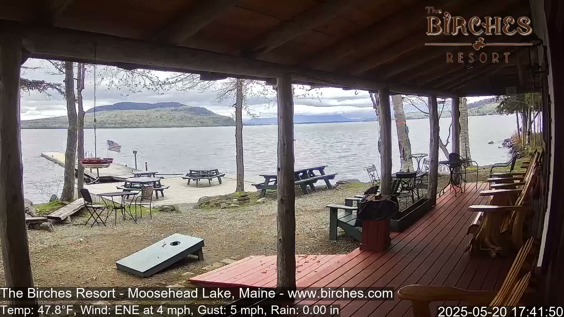 A lakeside porch with picnic tables and Adirondack chairs overlooks a calm lake under a partly cloudy sky.