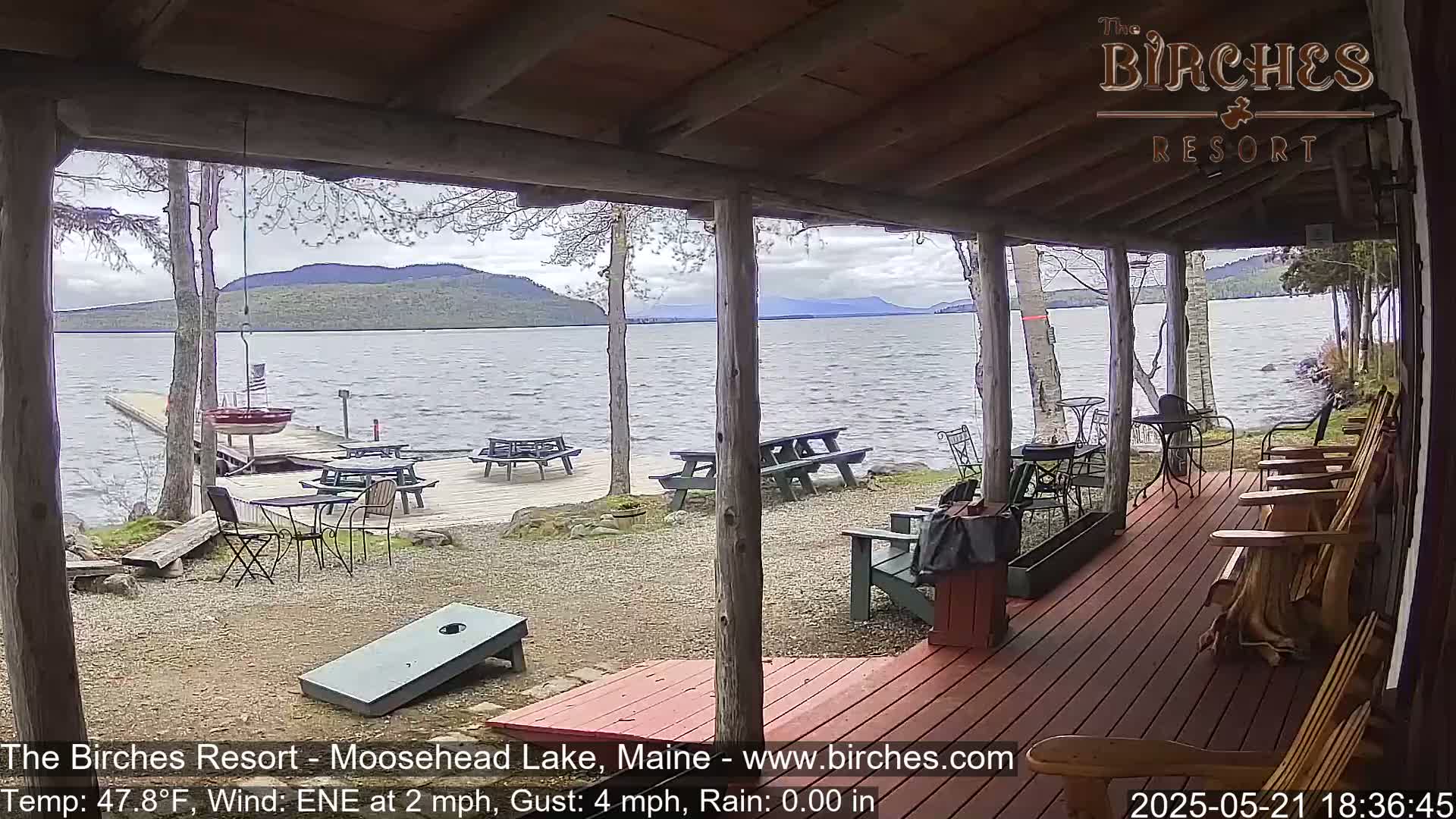A view from a covered porch shows a lake with a windy surface, picnic tables, Adirondack chairs, and a cornhole game set on a gravel area.