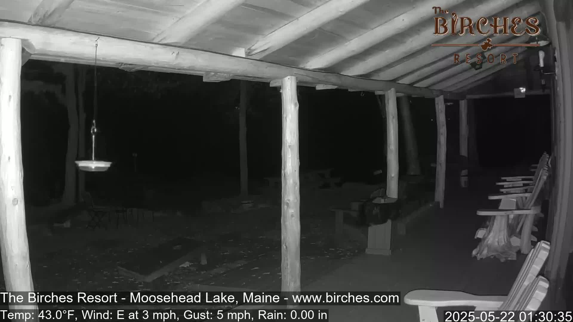 A nighttime, monochrome view of a covered porch with wooden posts, a bird feeder, and several empty Adirondack chairs, under calm, dry conditions.