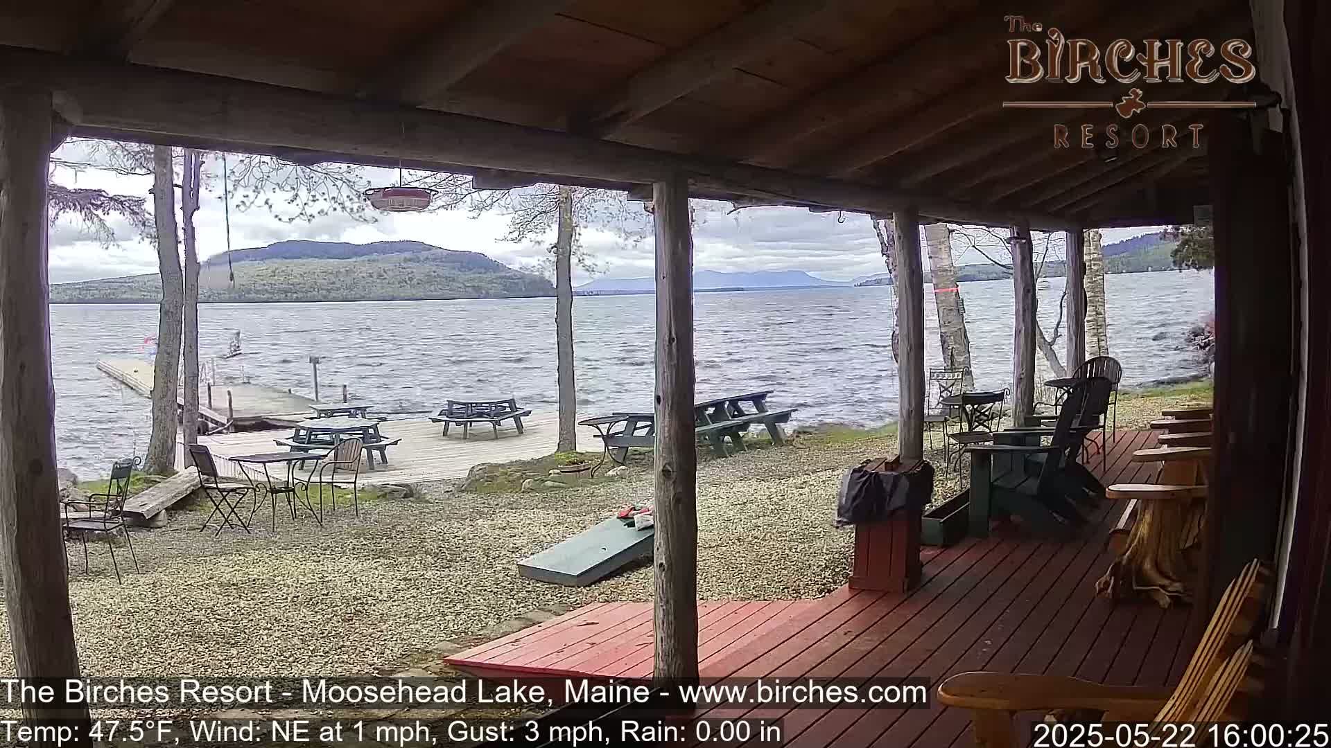 A lakeside porch with picnic tables and adirondack chairs overlooks a choppy lake under a cloudy sky.