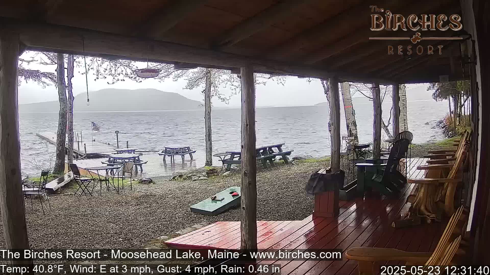 A rainy view from a lakeside porch shows several picnic tables and Adirondack chairs situated on a gravel area overlooking a choppy lake under a gray sky.