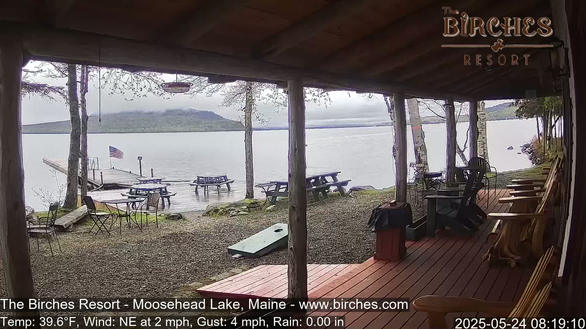 A lakeside porch with picnic tables and Adirondack chairs overlooks a calm lake under an overcast sky.