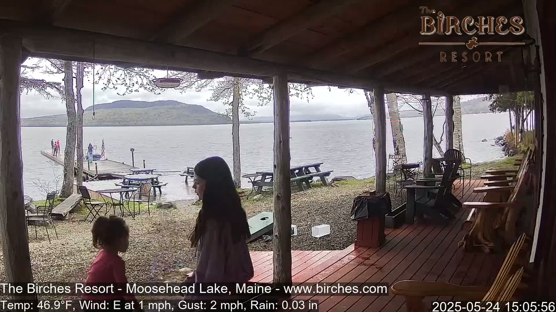 Two girls stand on a covered porch overlooking a lake on a rainy day, with picnic tables and Adirondack chairs visible on the deck.