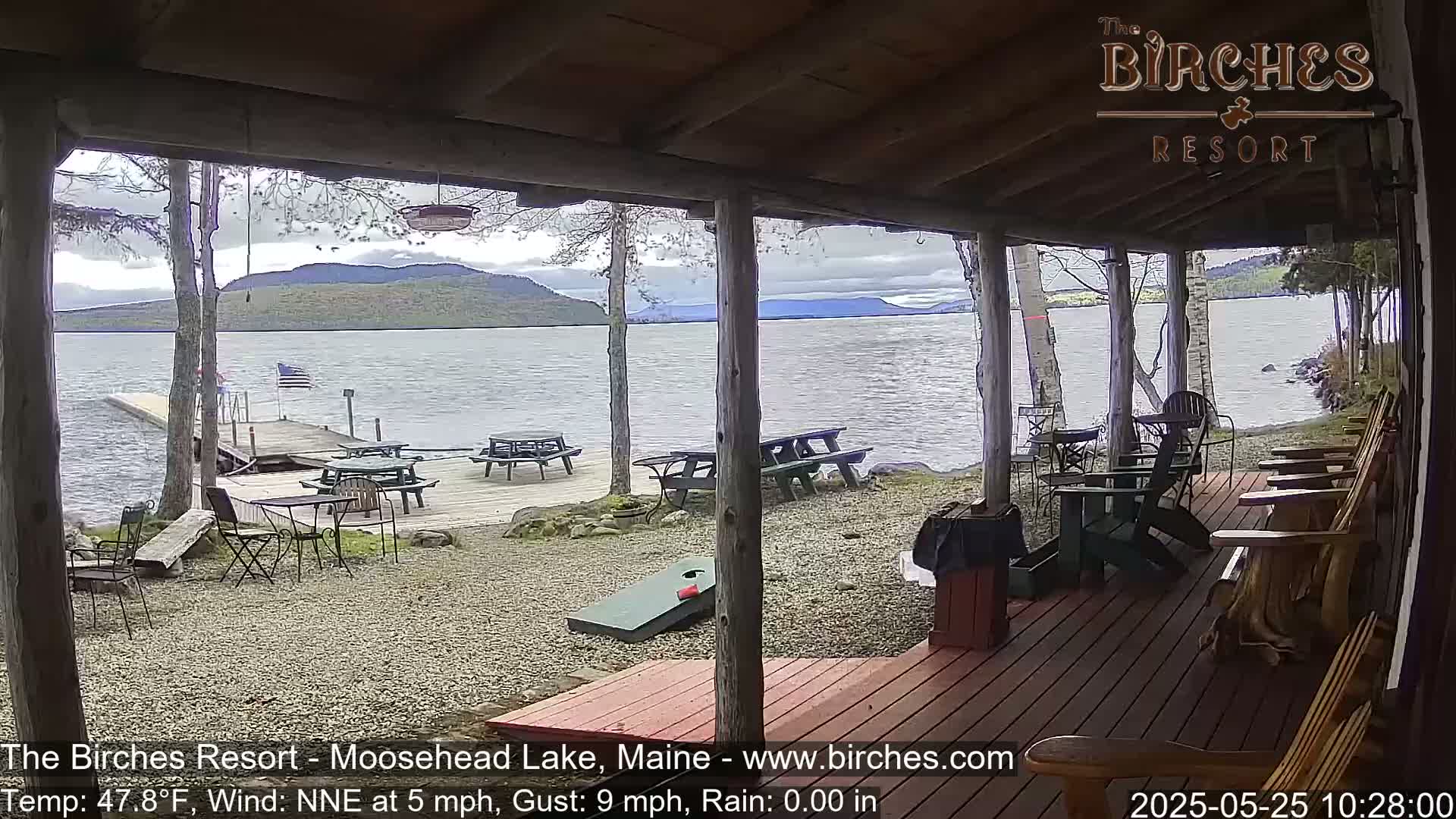 A lakeside porch with picnic tables and Adirondack chairs overlooks calm water and distant mountains under an overcast sky.