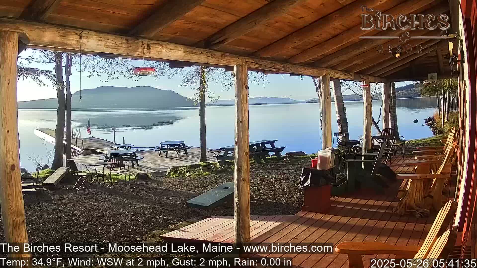 A wooden porch overlooks a calm lake with mountains in the distance under a clear, sunny sky.
