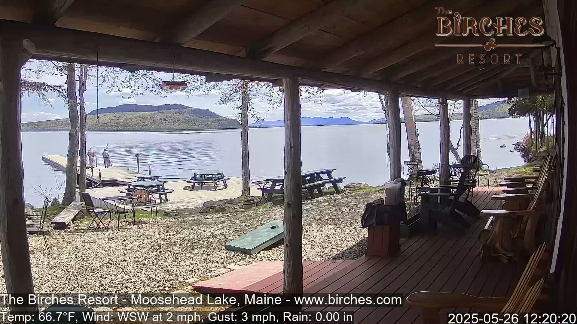 A lakeside porch with picnic tables and Adirondack chairs overlooks a calm lake and distant mountains under a partly cloudy sky.