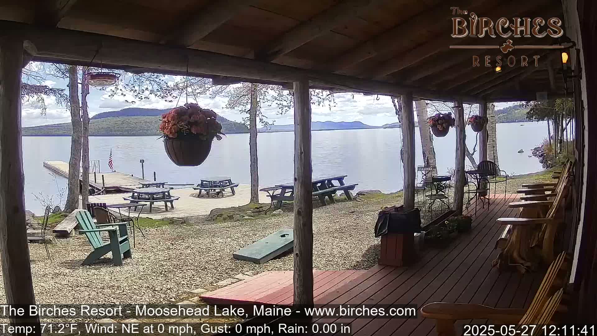 A covered porch overlooks a calm lake with picnic tables and Adirondack chairs on a partly cloudy day.