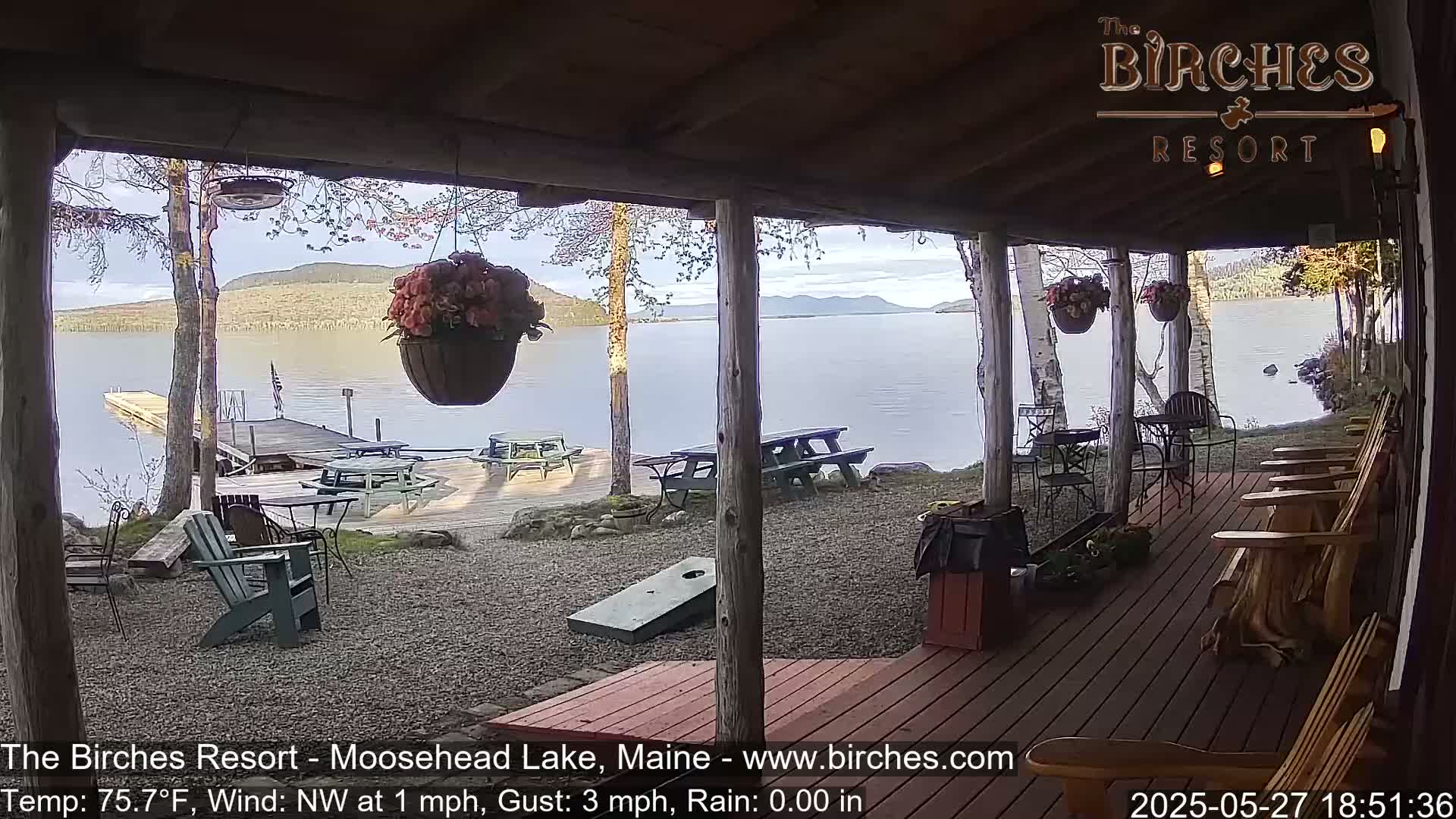 A lakeside porch with wooden rocking chairs and picnic tables overlooks a calm lake on a partly sunny day.