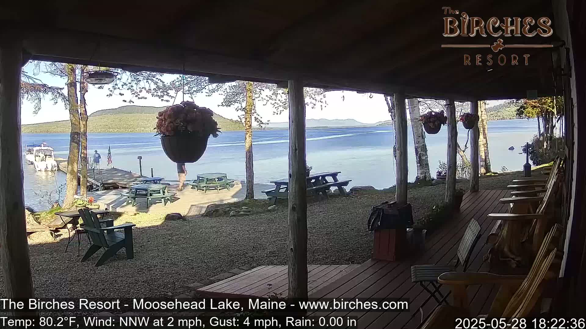A lakeside porch with picnic tables and rocking chairs overlooks a calm lake and distant mountains on a sunny day with a gentle northwest wind.